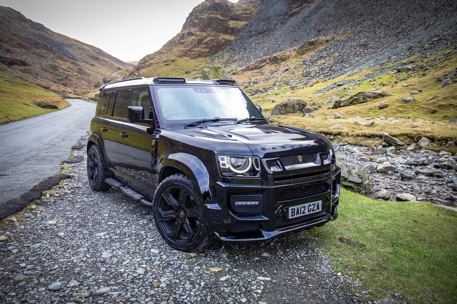 Front quarter view of a custom wrapped Land Rover Defender 110 L663 2020+. Shot outside near mountains. Fitted with Barugzai Sabre 22” alloy wheels, in Gloss Black.