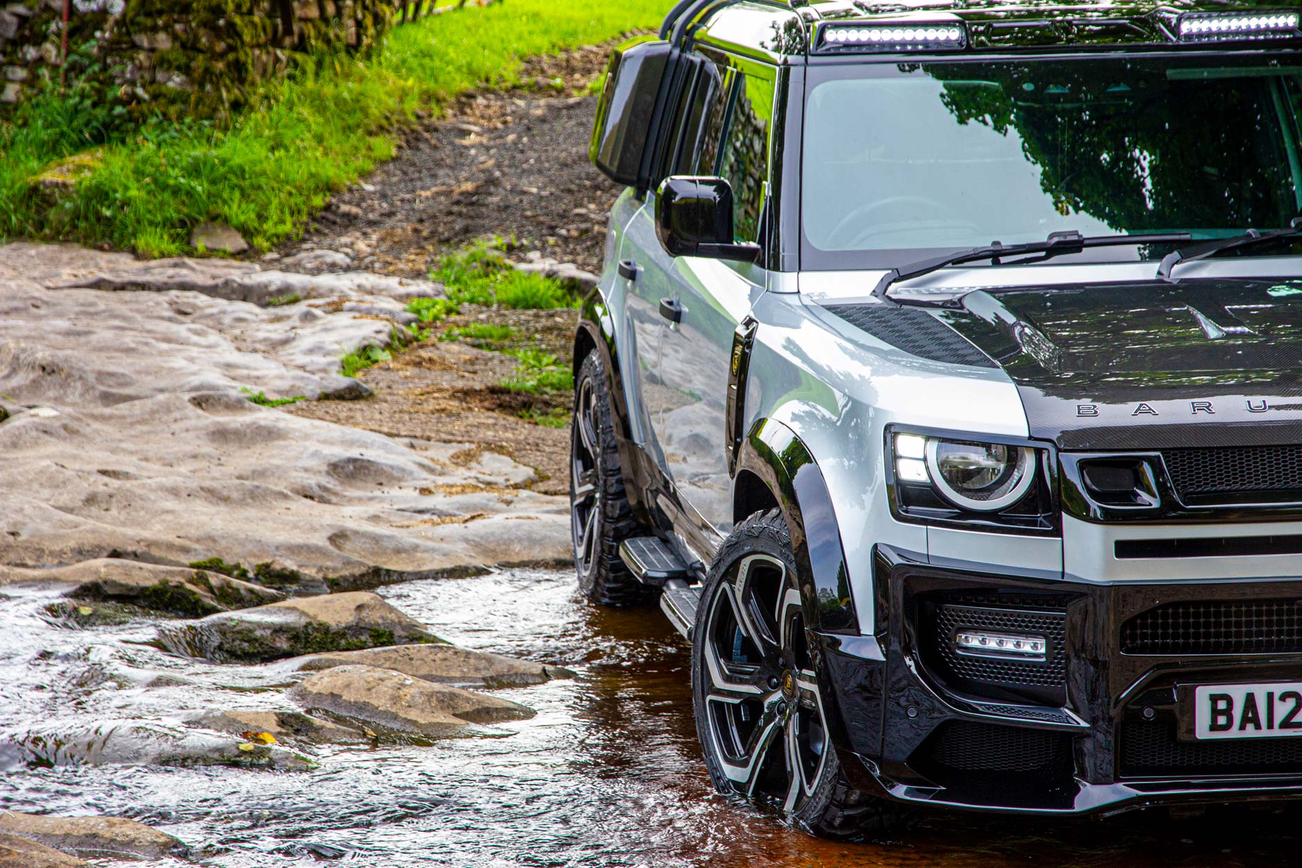 Front quarter detail view of a Silver Land Rover Defender 110 L663 2020+. Shot outside a hiking trail. Fitted with Barugzai Sabre 22” alloy wheels, in Gloss Black Polished Face.
