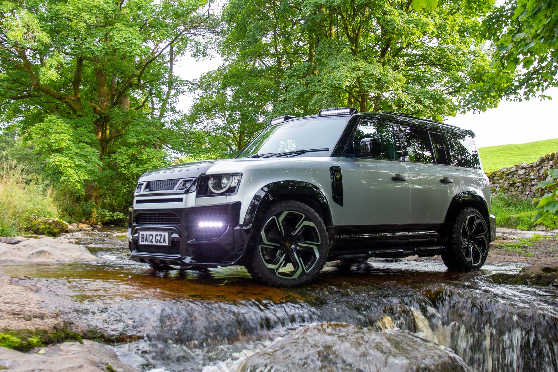 Front quarter view of a Silver Land Rover Defender 110 L663 2020+. Shot outside a hiking trail. Fitted with Barugzai Sabre 22” alloy wheels, in Gloss Black Polished Face.