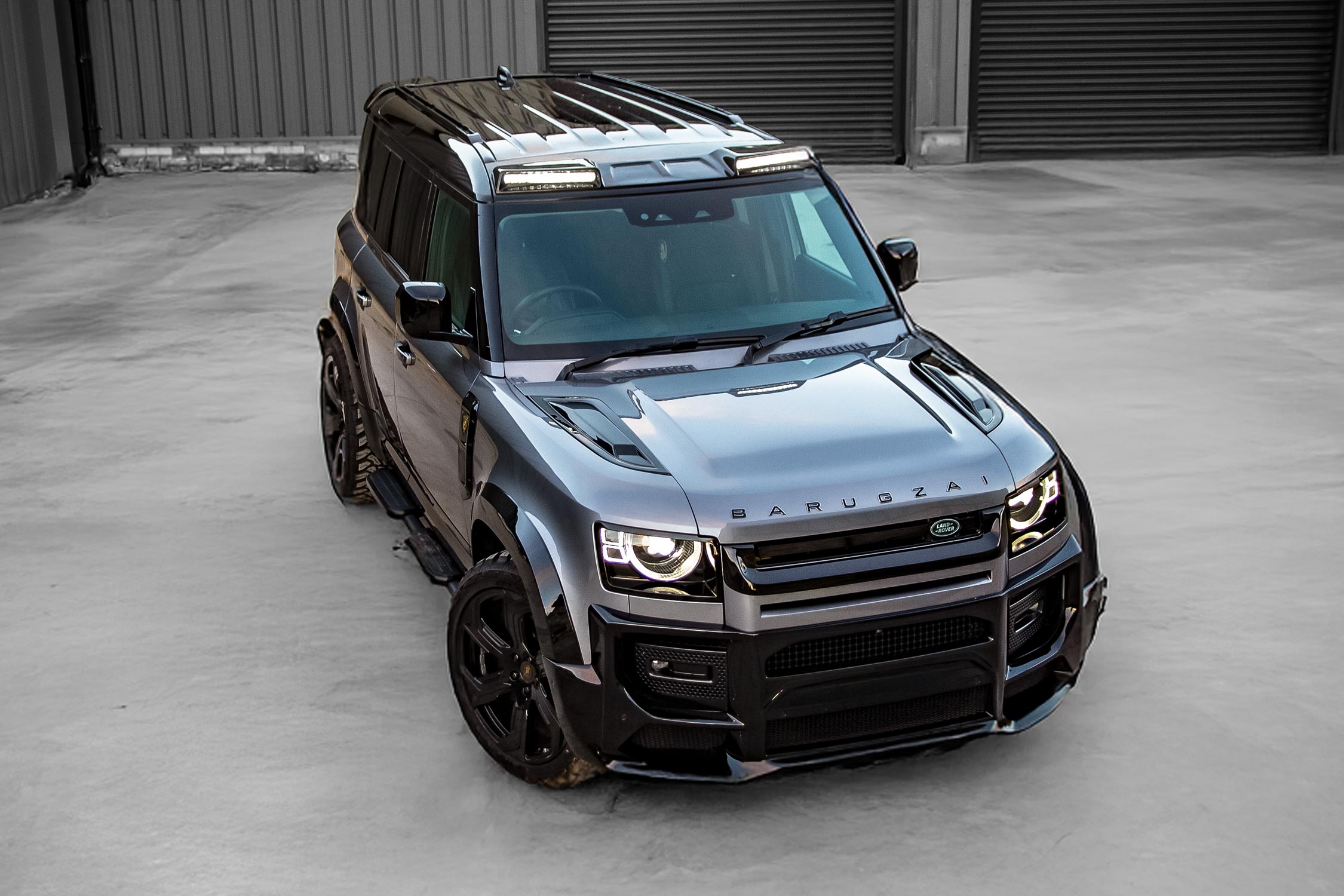 Front quarter view of a silver Land Rover Defender 110 L663 2020+. Shot outside an empty warehouse. Fitted with Barugzai Sabre 22” alloy wheels, in Gloss Black.