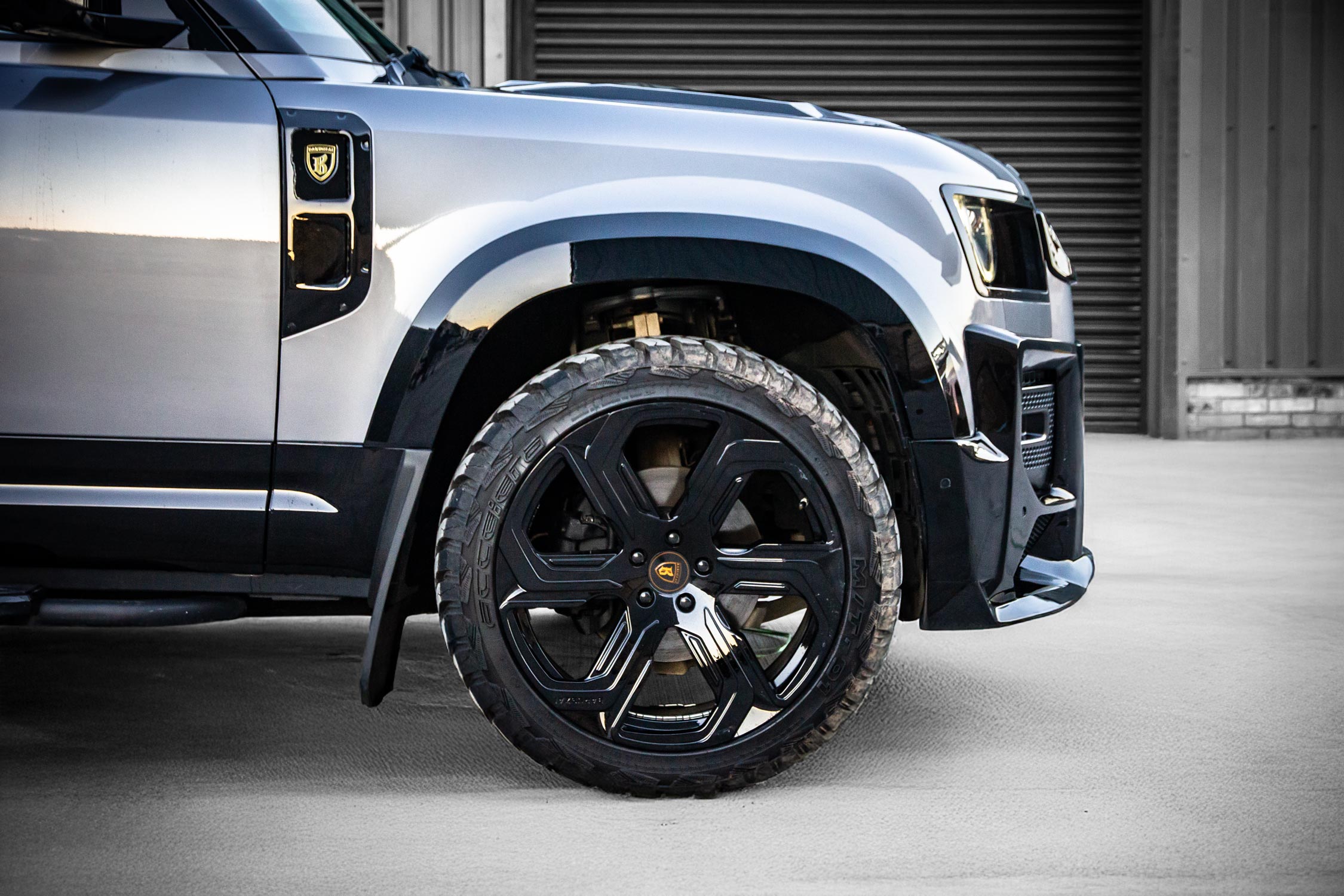 Side detail view of a silver Land Rover Defender 110 L663 2020+. Shot outside an empty warehouse. Fitted with Barugzai Sabre 22” alloy wheels, in Gloss Black.
