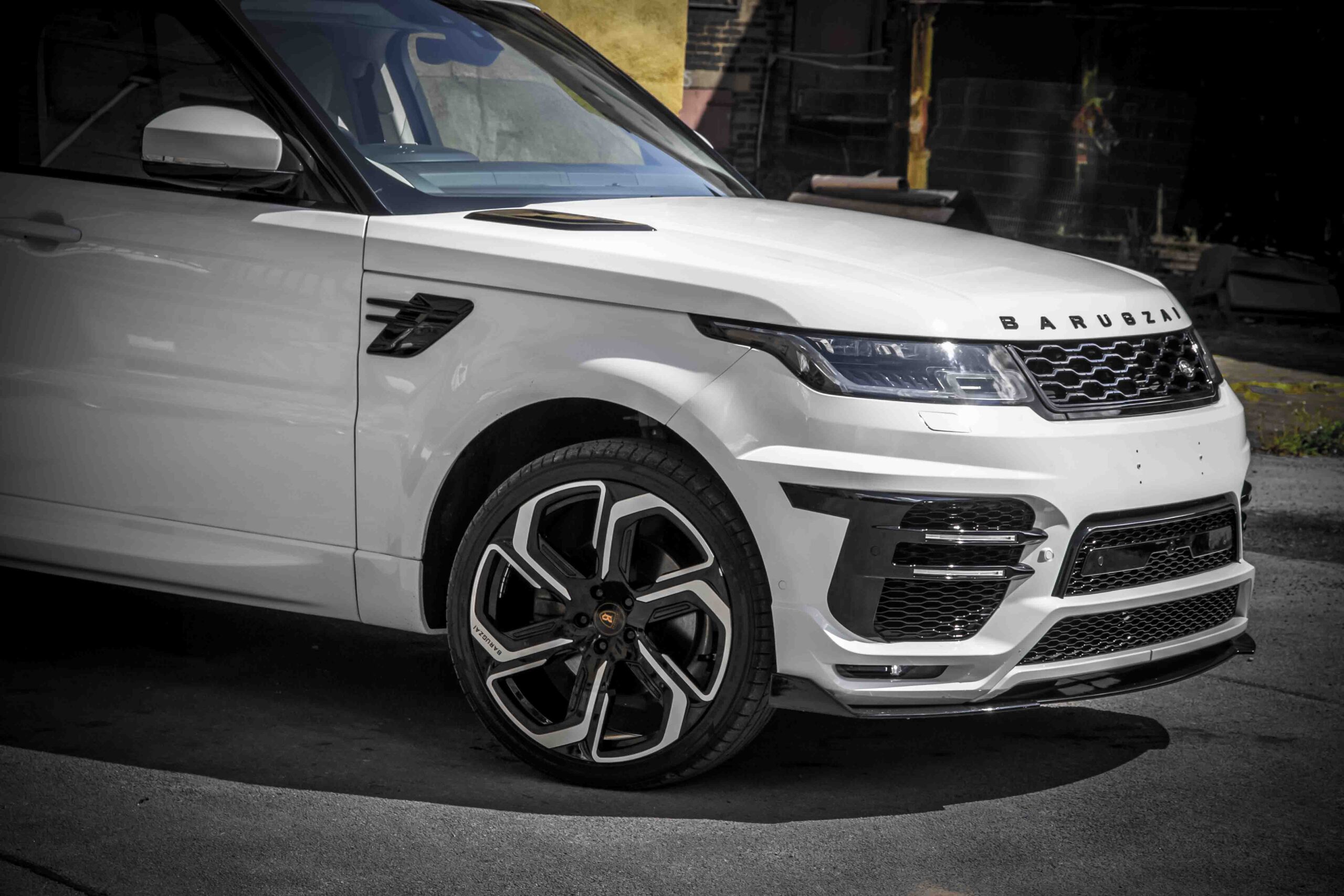 Front quarter detail view of a White Range Rover Sport L494 2018-2022. Shot outside an old mill. Fitted with Barugzai Sabre 22” alloy wheels, in Gloss Black Polished Face.