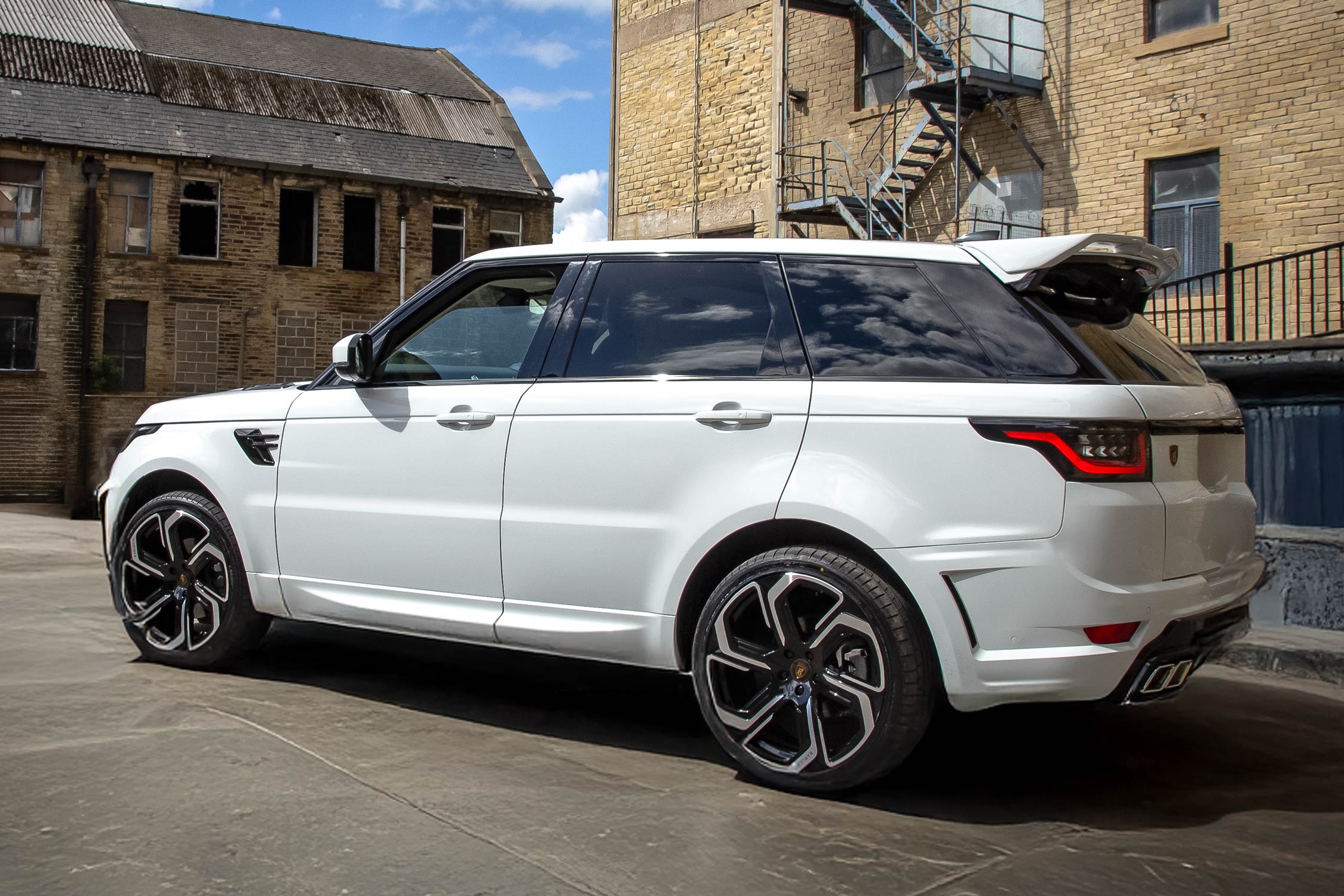 Rear quarter view of a White Range Rover Sport L494 2018-2022. Shot outside an old mill. Fitted with Barugzai Sabre 22” alloy wheels, in Gloss Black Polished Face.