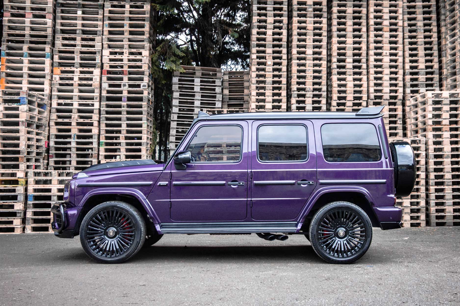 Side view of a Purple Mercedes G Wagon 2018+. Shot outside a yard full of pallets. Fitted with Barugzai Casanova 22/23” alloy wheels, in Gloss Black.
