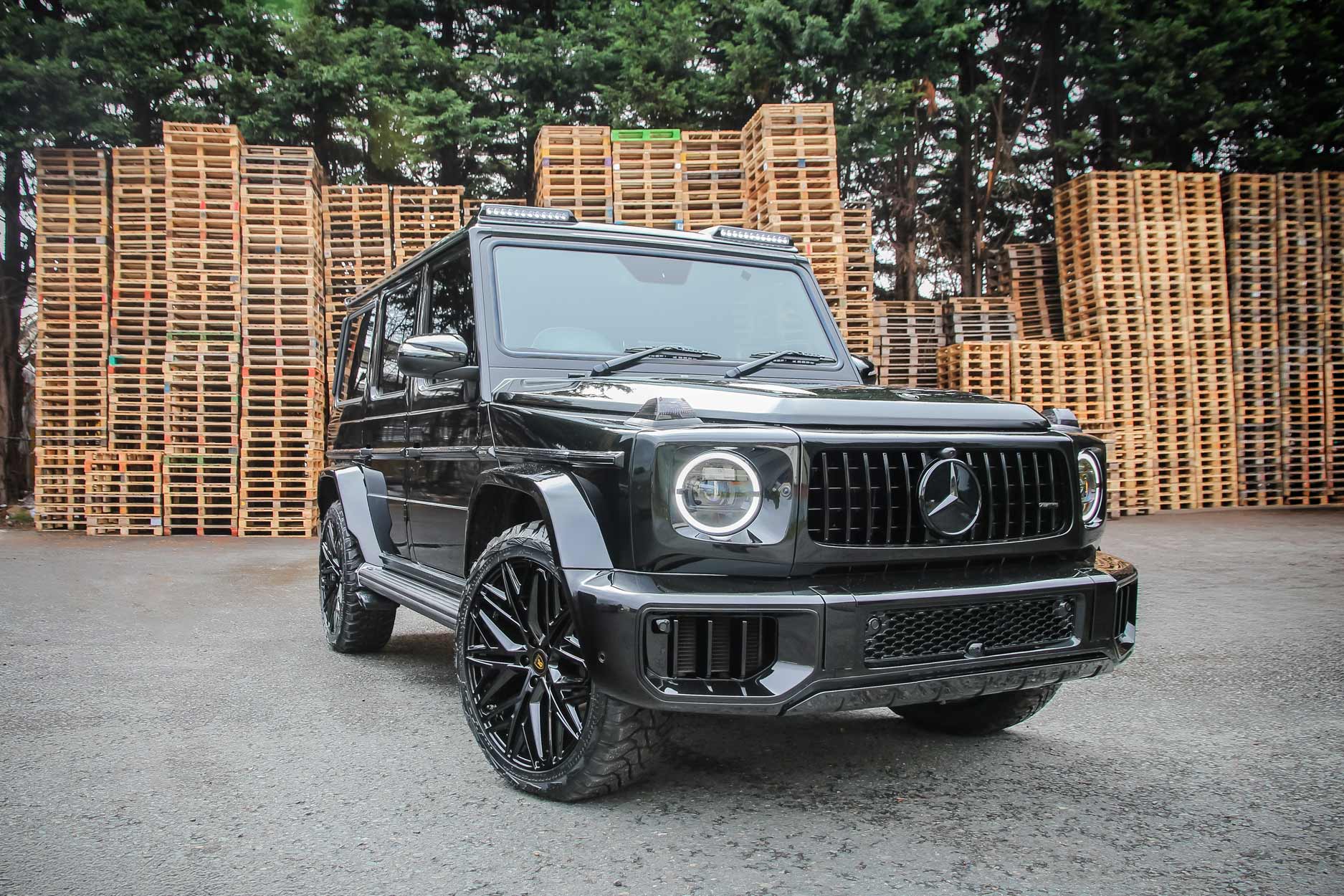 Front quarter view of a Black Mercedes G Wagon facelifted to 2025 model. Parked outside a pallet yard. Fitted with Barugzai Shuracan 22” alloy wheels, in Gloss Black.