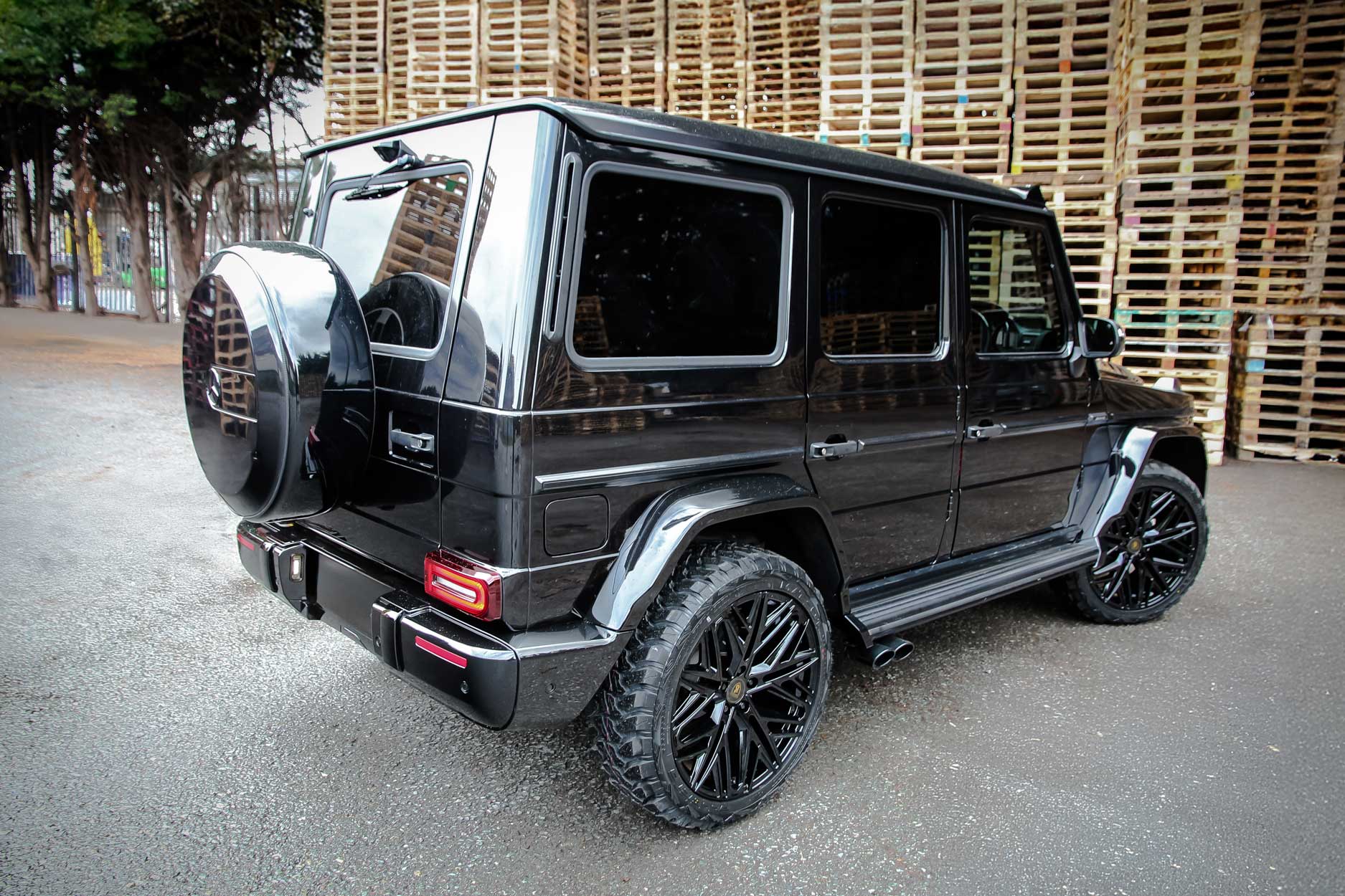 Rear quarter view of a Black Mercedes G Wagon facelifted to 2025 model. Parked outside a pallet yard. Fitted with Barugzai Shuracan 22” alloy wheels, in Gloss Black.