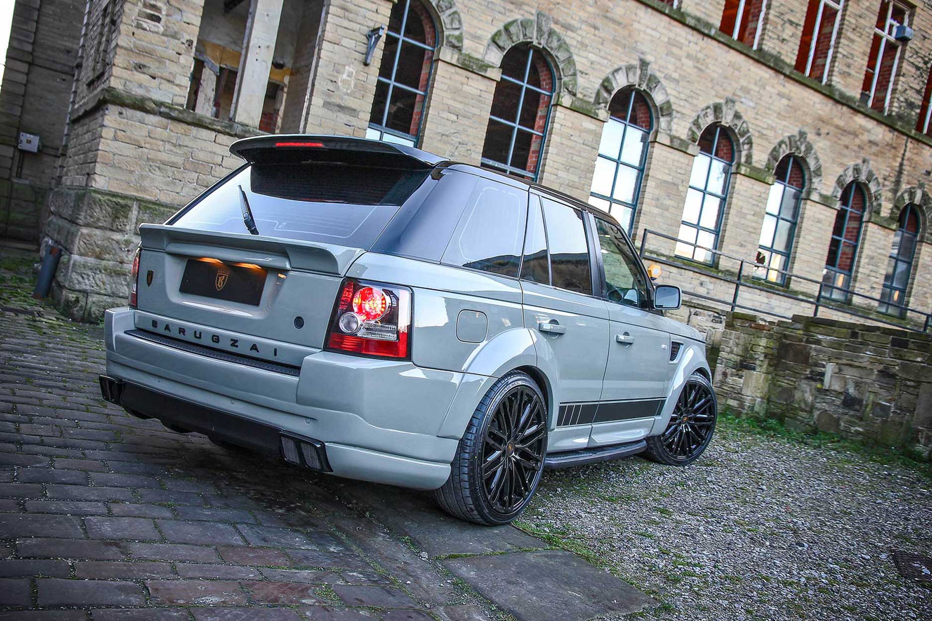 Rear quarter view of a Nardo Grey Range Rover Sport L320 2009-2012. Shot outside an old mill. Fitted with Barugzai Dawn 22” alloy wheels, in Gloss Black.