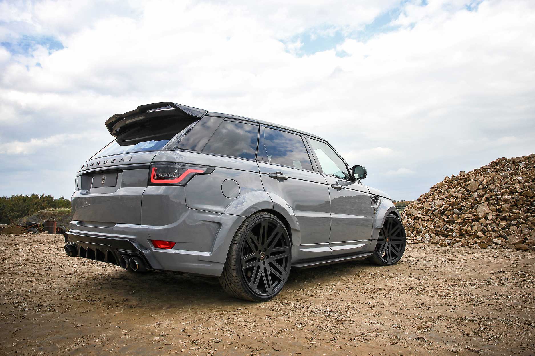 Rear quarter view of a Nardo Grey Range Rover Sport L494 2013-2017. Shot outside a quarry. Fitted with Barugzai EXEL 24” alloy wheels, in Gloss Black.