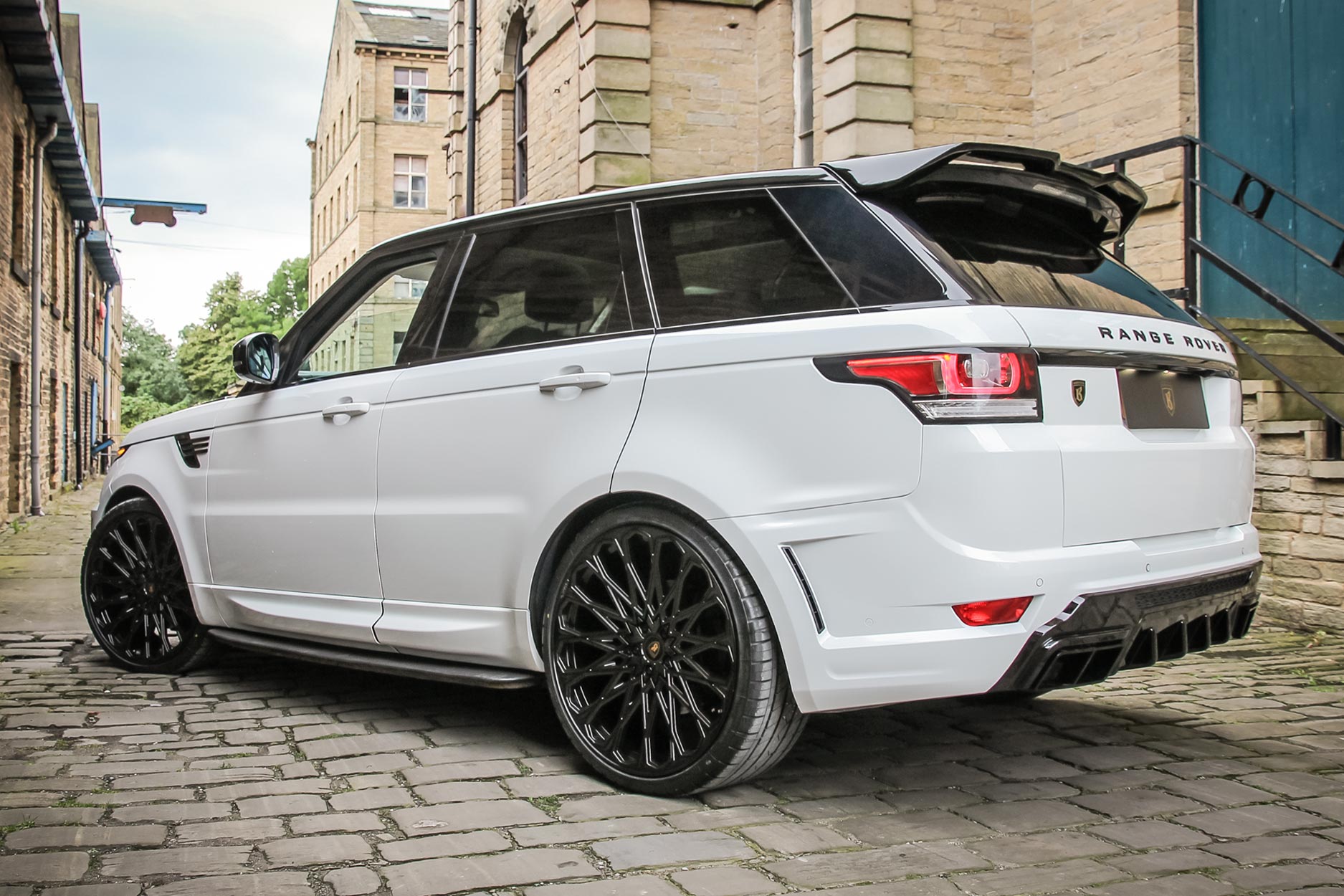 Rear quarter view of a White Range Rover Sport L494 2013-2017. Shot outside an old mill. Fitted with Barugzai Majestic 24” alloy wheels, in Gloss Black.
