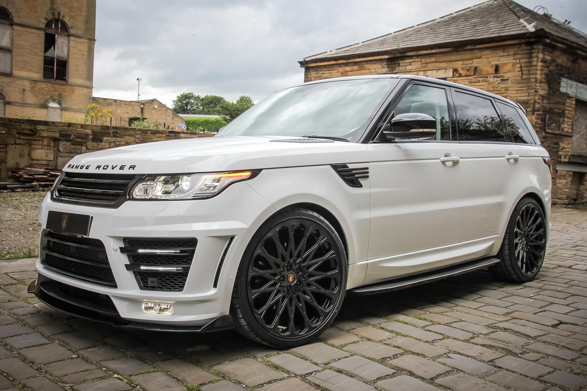 Front quarter view of a White Range Rover Sport L494 2013-2017. Shot outside an old mill. Fitted with Barugzai Majestic 24” alloy wheels, in Gloss Black.