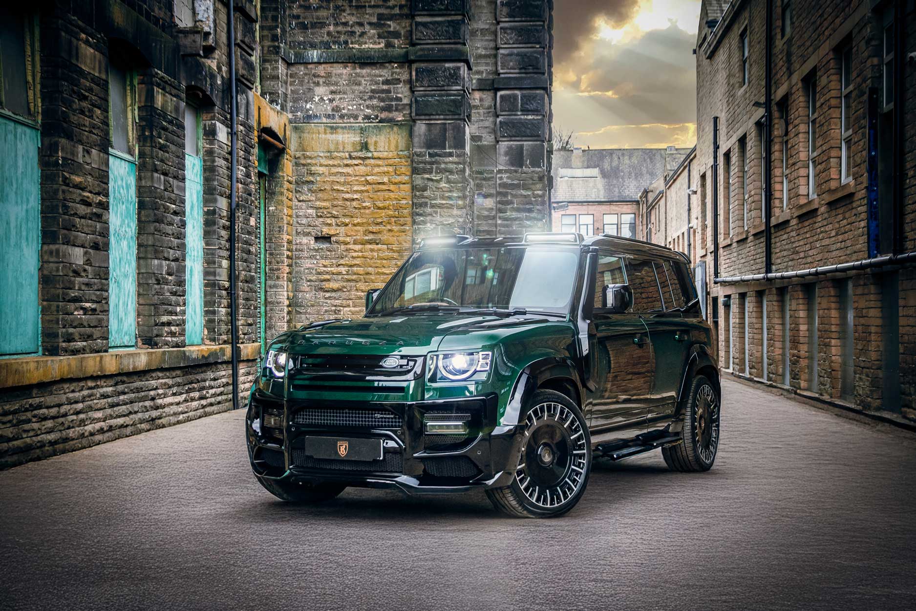 Front quarter view of a Green Land Rover Defender 110 L663 2020+. Shot outside an old mill. Fitted with Barugzai Monoblok 23” alloy wheels, in Gloss Black Polished Face.