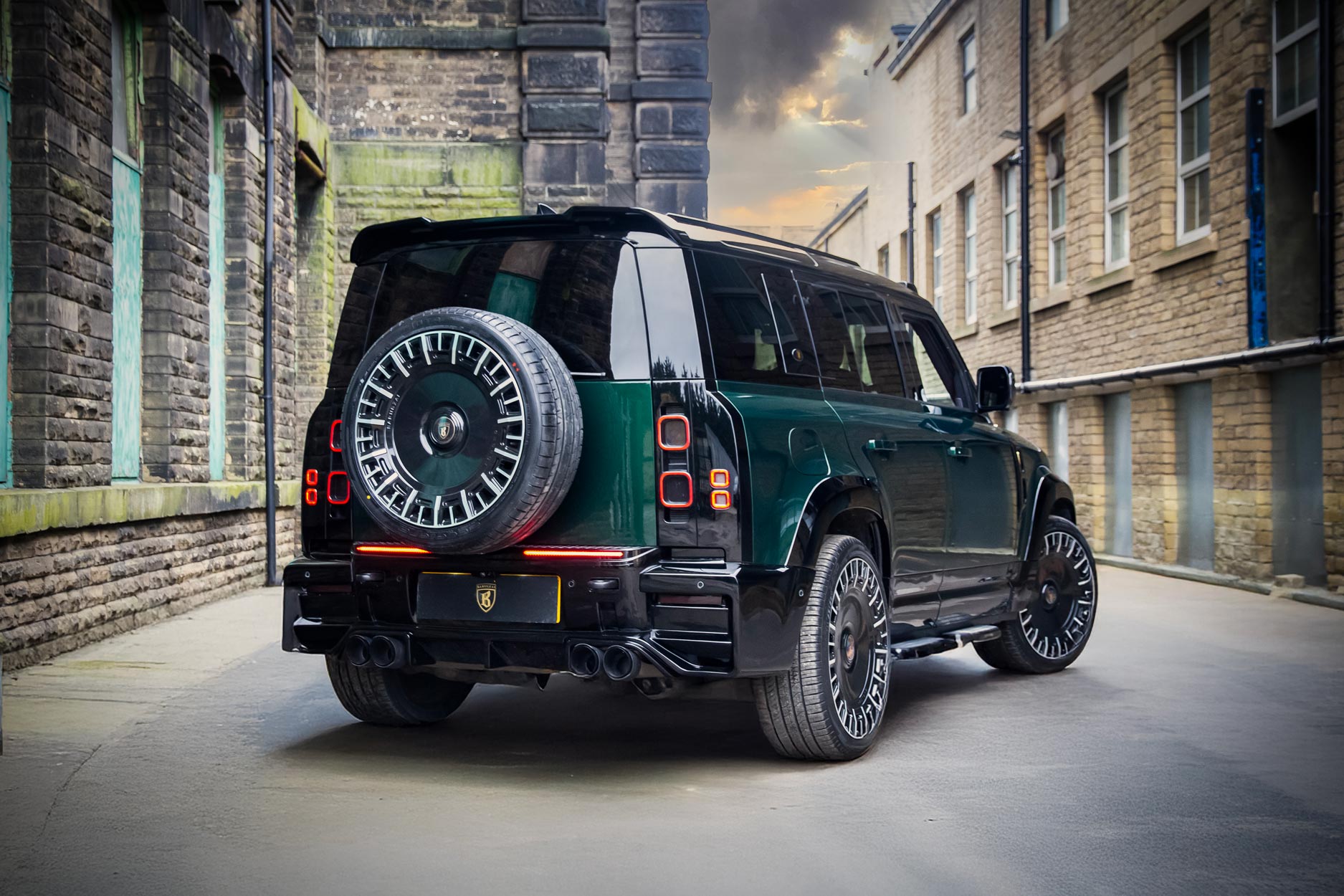 Rear quarter view of a Green Land Rover Defender 110 L663 2020+. Shot outside an old mill. Fitted with Barugzai Monoblok 23” alloy wheels, in Gloss Black Polished Face.