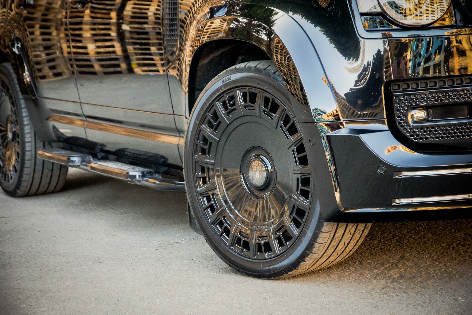 Front quarter detail view of a Black Land Rover Defender 110 L663 2020+. Shot outside pallet warehouse. Fitted with Barugzai Monoblok 23” alloy wheels, in Gloss Black.
