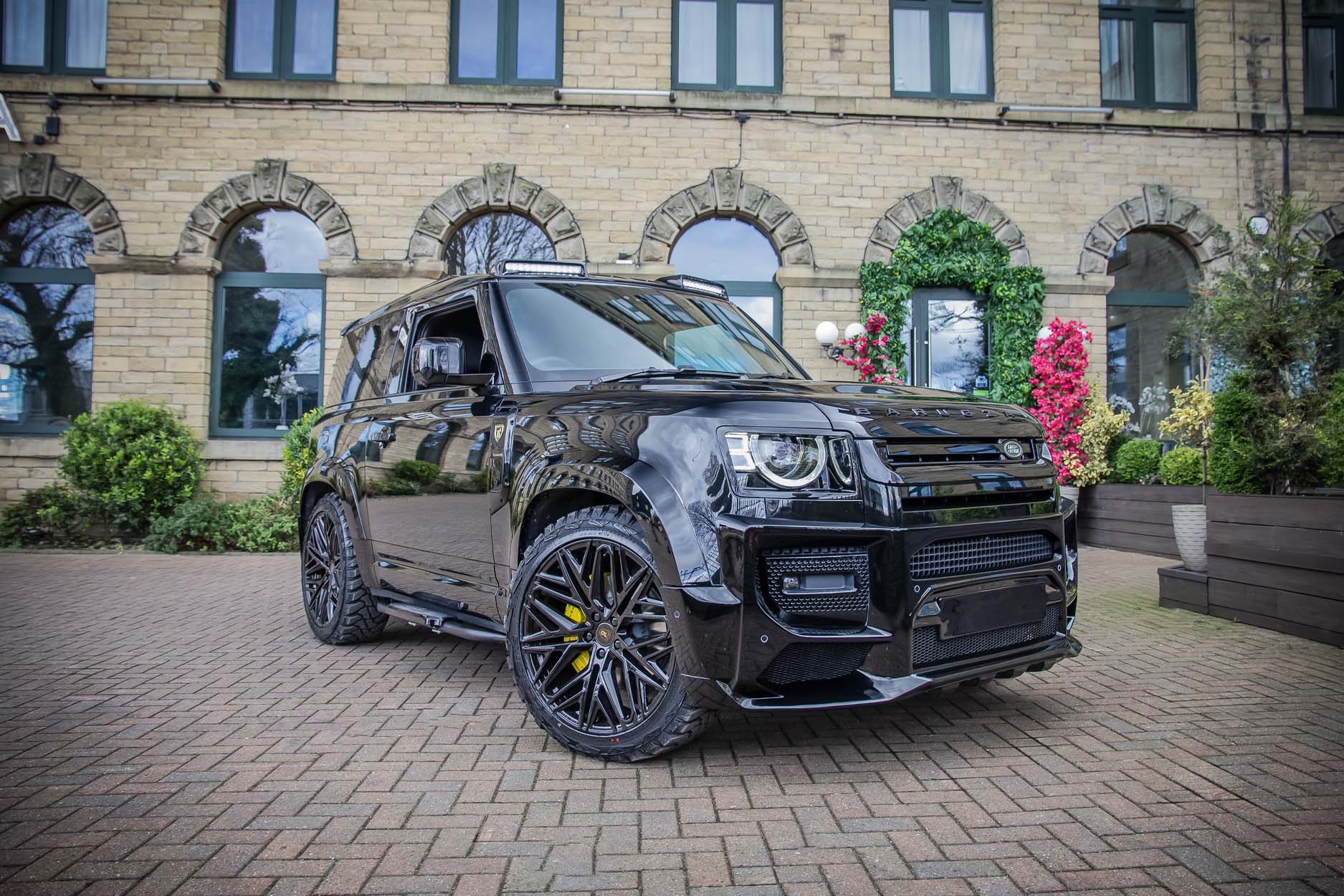 Front quarter view of a Black Land Rover Defender 90 L663. Parked outside a restaurant Fitted with Barugzai Shuracan 22” alloy wheels, in Gloss Black.