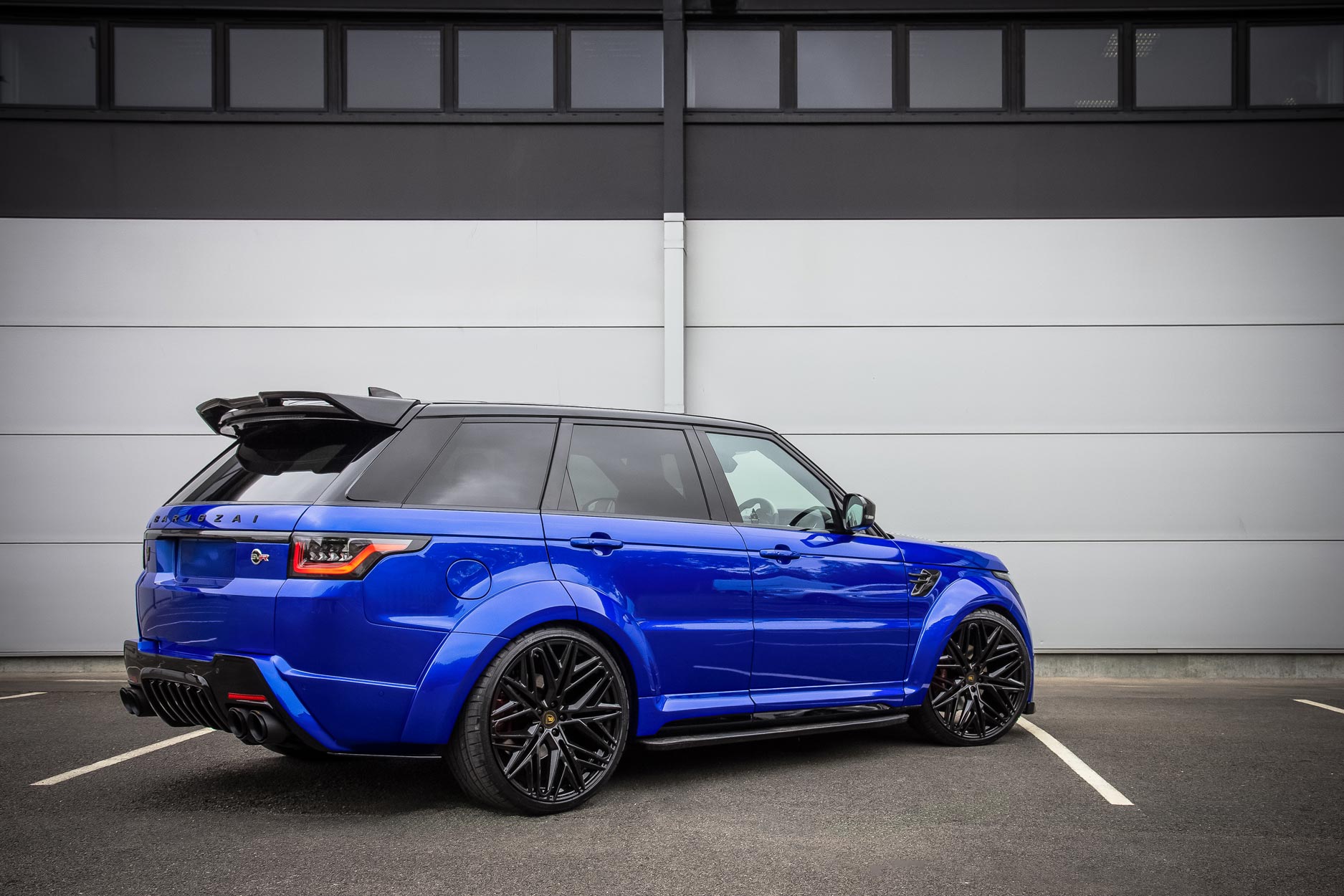 Rear quarter view of a Blue Range Rover Sport L494 2018-2022. Parked on a road with a grey background. Fitted with Barugzai Shuracan 24” alloy wheels, in Gloss Black.