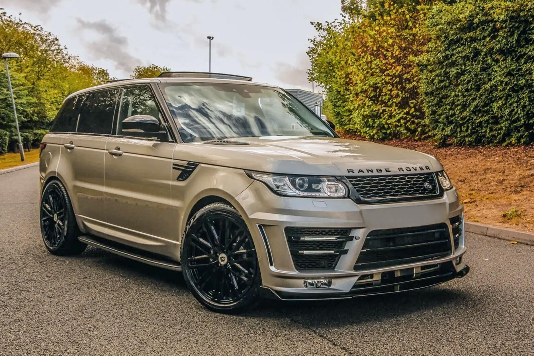 Front quarter view of a Champagne Range Rover Sport L494 2013-2017. Shot outside with a scenic setting. Fitted with Barugzai Dawn 22” alloy wheels, in Gloss Black.