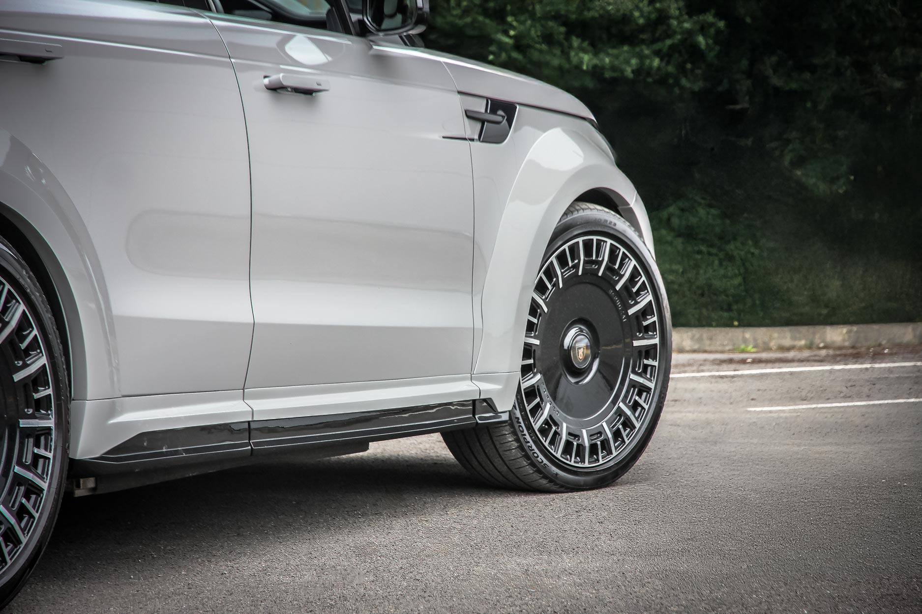 Side quarter detail view of a Nardo Grey Range Rover Sport L461 2023+. Shot outside a scenic backdrop. Fitted with Barugzai Monoblok 23” alloy wheels, in Gloss Black Polished Face.