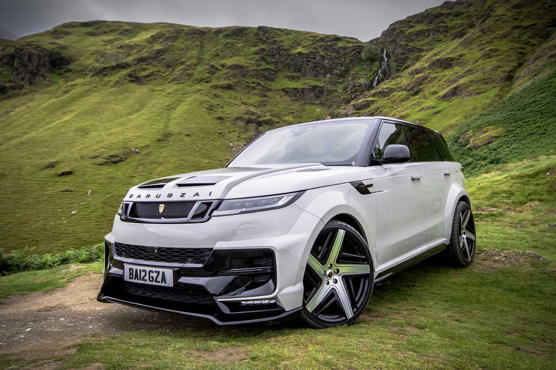 Front quarter view of a Nardo Grey Range Rover Sport 2023+. Parked outside a mountain scenery. Fitted with Barugzai Eleven 24” alloy wheels, in Gloss Black Polished Face.