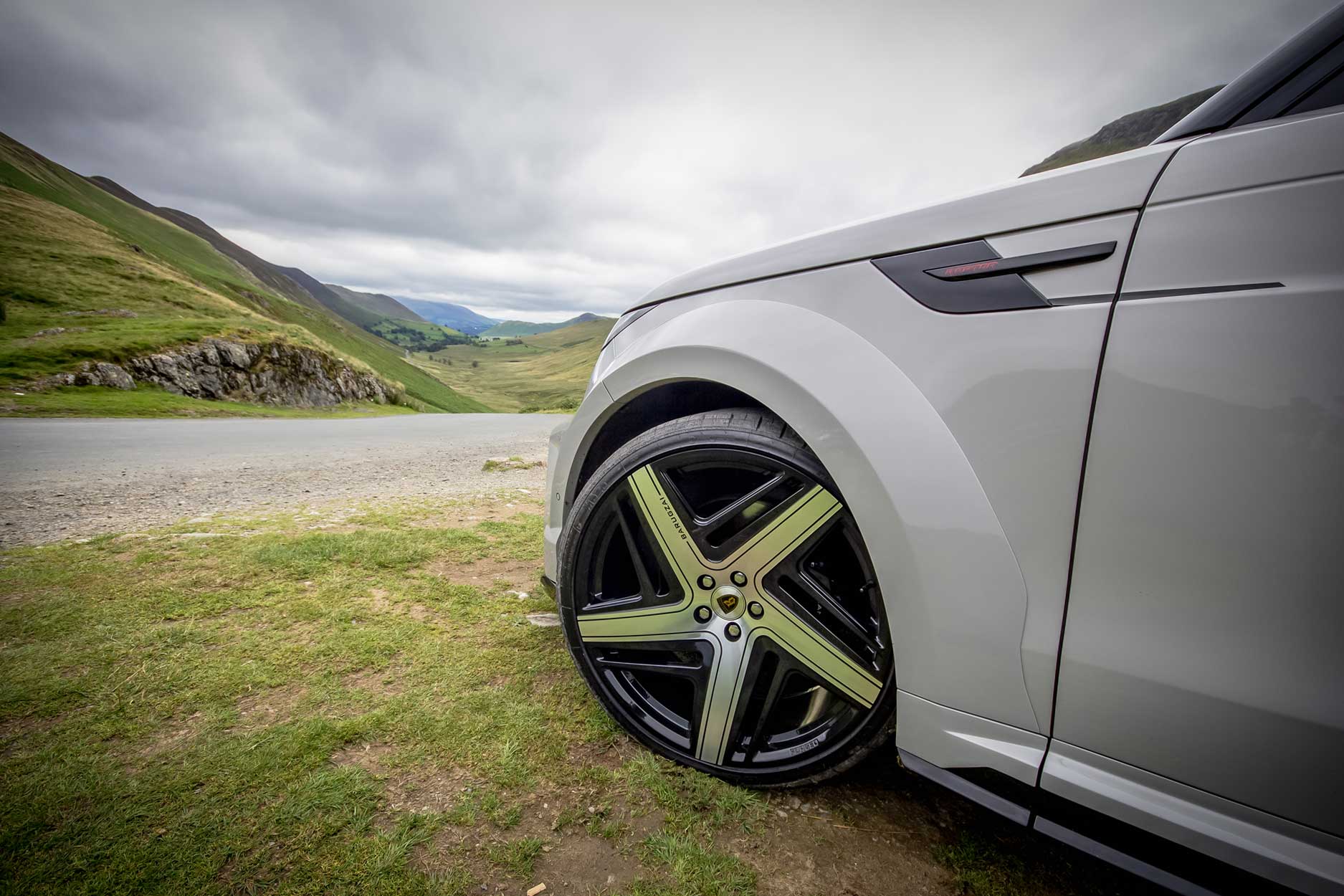 Side quarter detail view of a Nardo Grey Range Rover Sport 2023+. Parked outside a mountain scenery. Fitted with Barugzai Eleven 24” alloy wheels, in Gloss Black Polished Face.