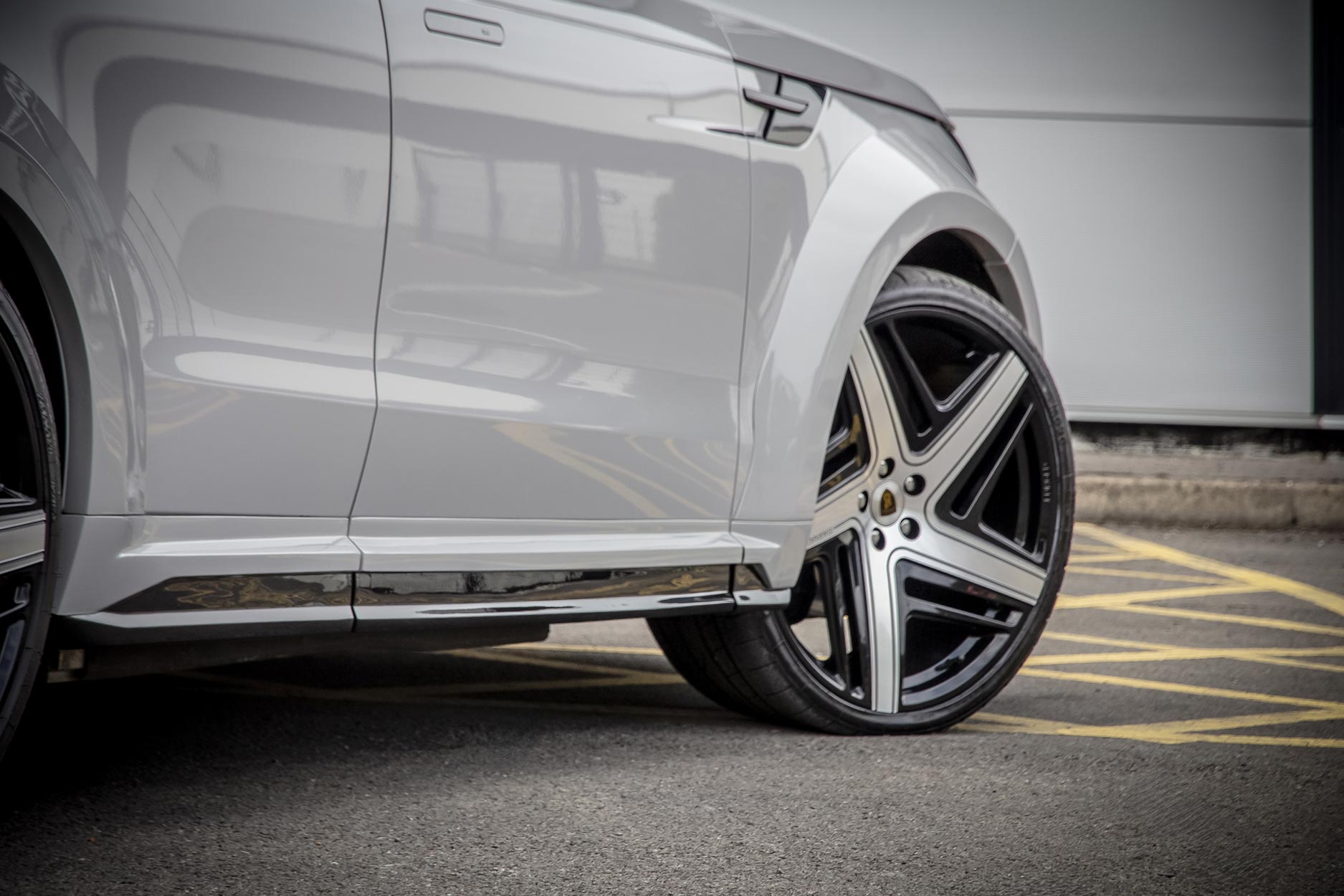 Side quarter detail view of a Nardo Grey Range Rover Sport 2023+. Parked on a road with a grey background. Fitted with Barugzai Eleven 24” alloy wheels, in Gloss Black Polished Face.
