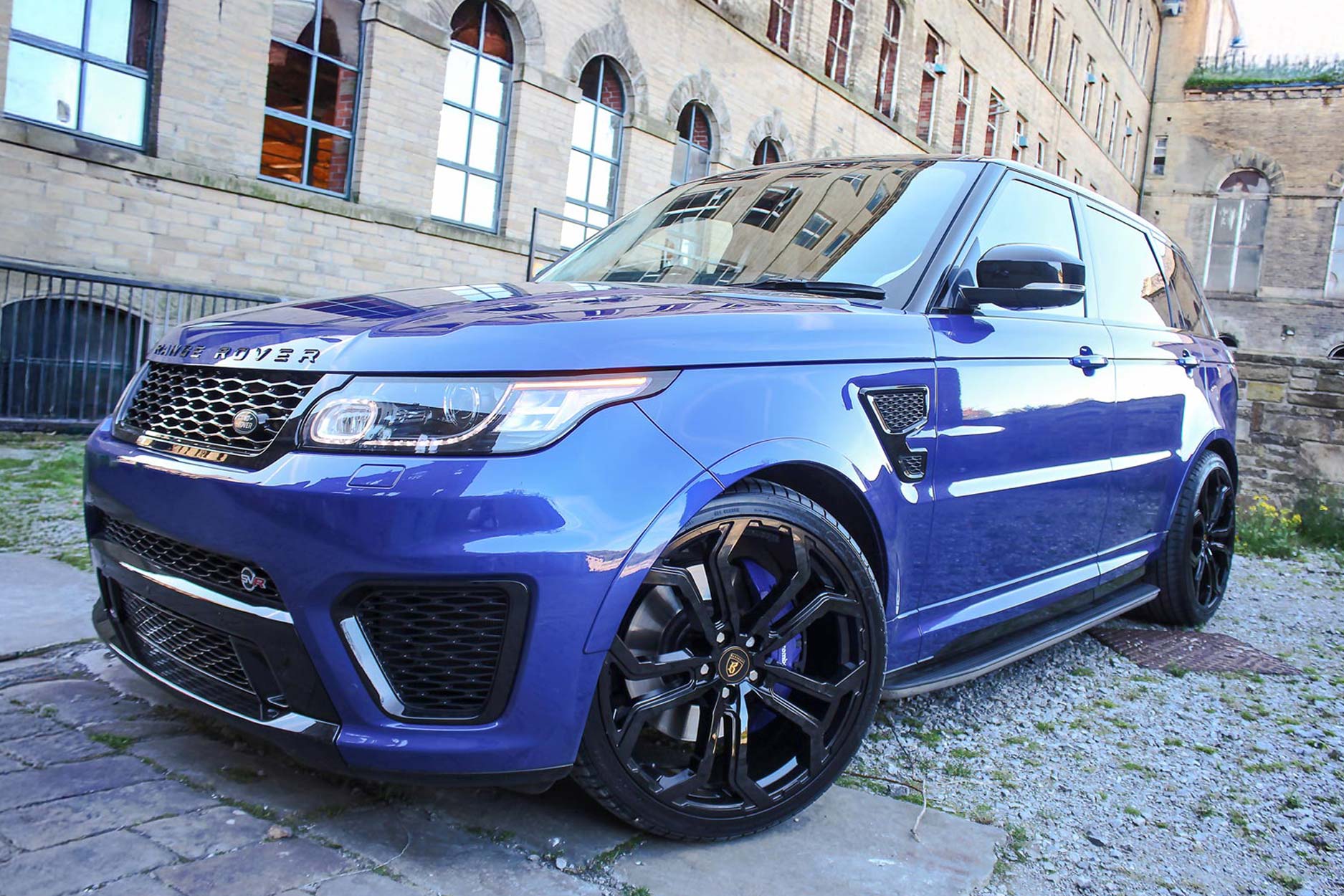 Front quarter view of a Blue Range Rover Sport SVR L494 2013-2017. Shot outside an old mill. Fitted with Barugzai Opium 22” alloy wheels, in Gloss Black.